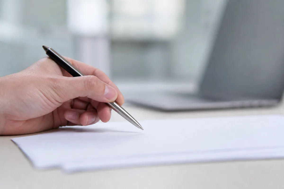 Main tenant un stylo au-dessus d’une liasse de papiers avec un ordinateur portable en arrière-plan flouté, ambiance bureau lumineux